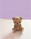 A small beige teddy bear ornament sitting on a white surface against a lavender-purple background.