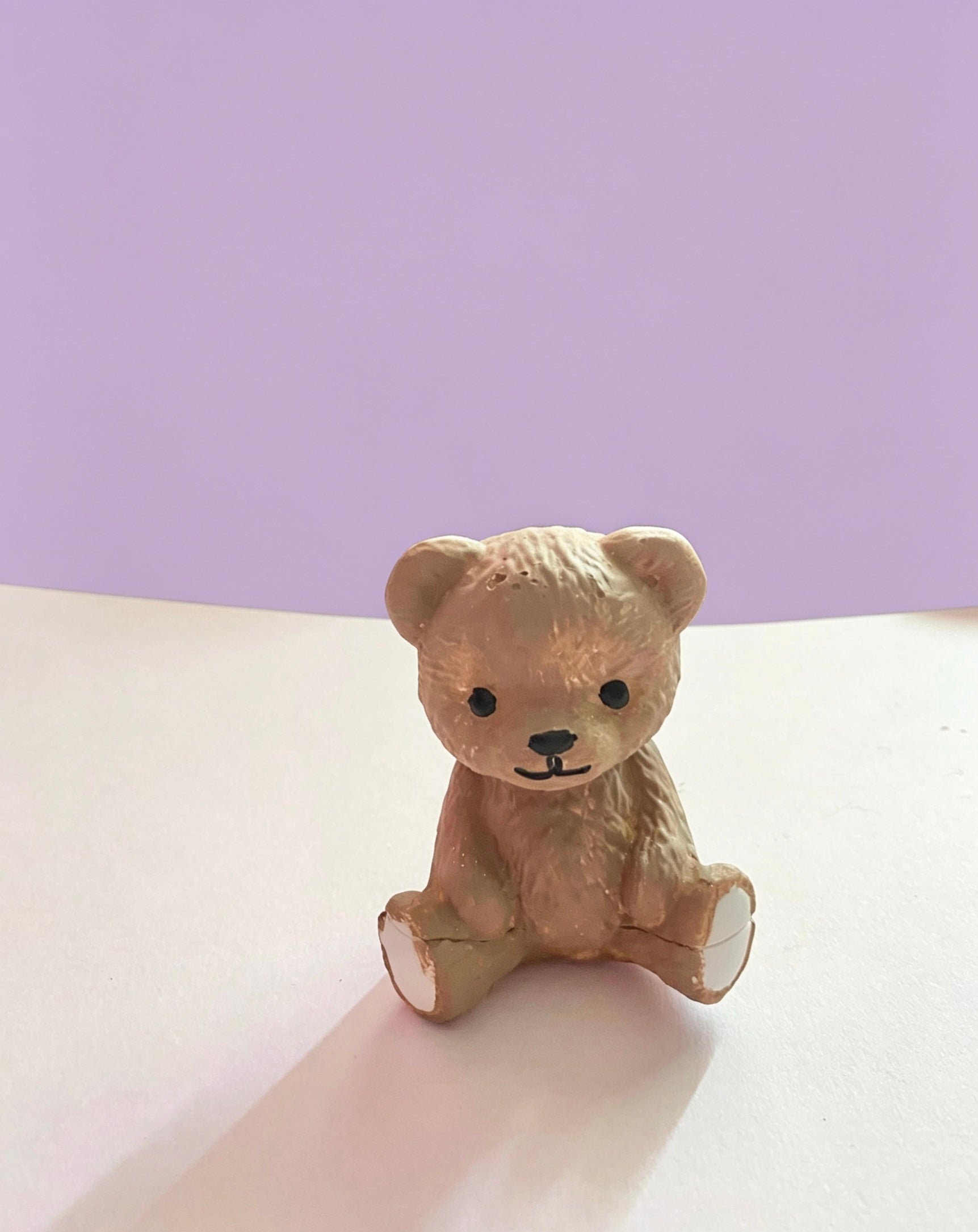 A small beige teddy bear ornament sitting on a white surface against a lavender-purple background.
