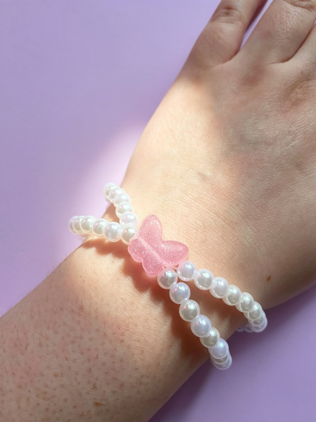 Pearl bracelet with a pink butterfly charm on a wrist against a light purple background