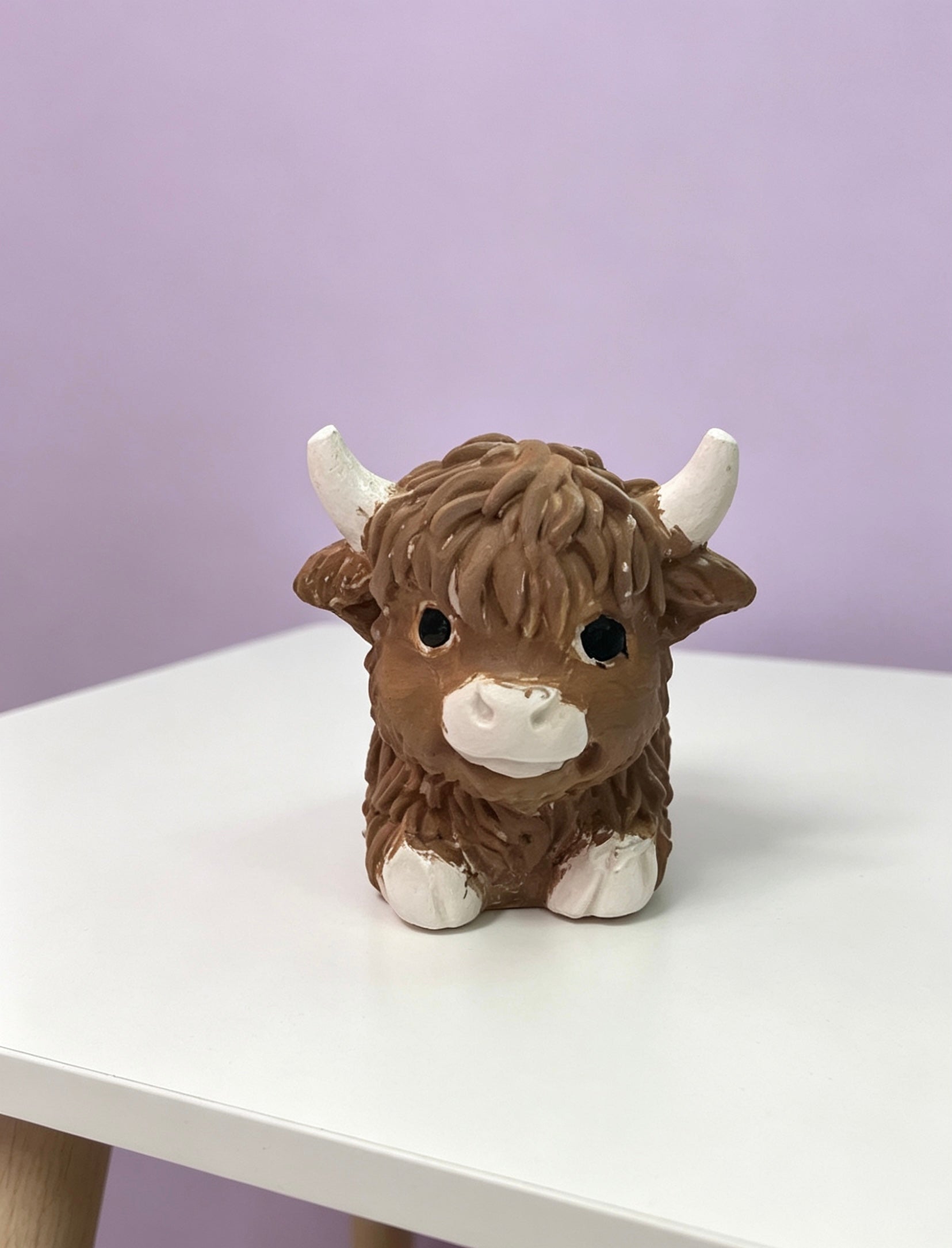 A small brown and white Highland cow ornament sitting on a white table against a light purple background.
