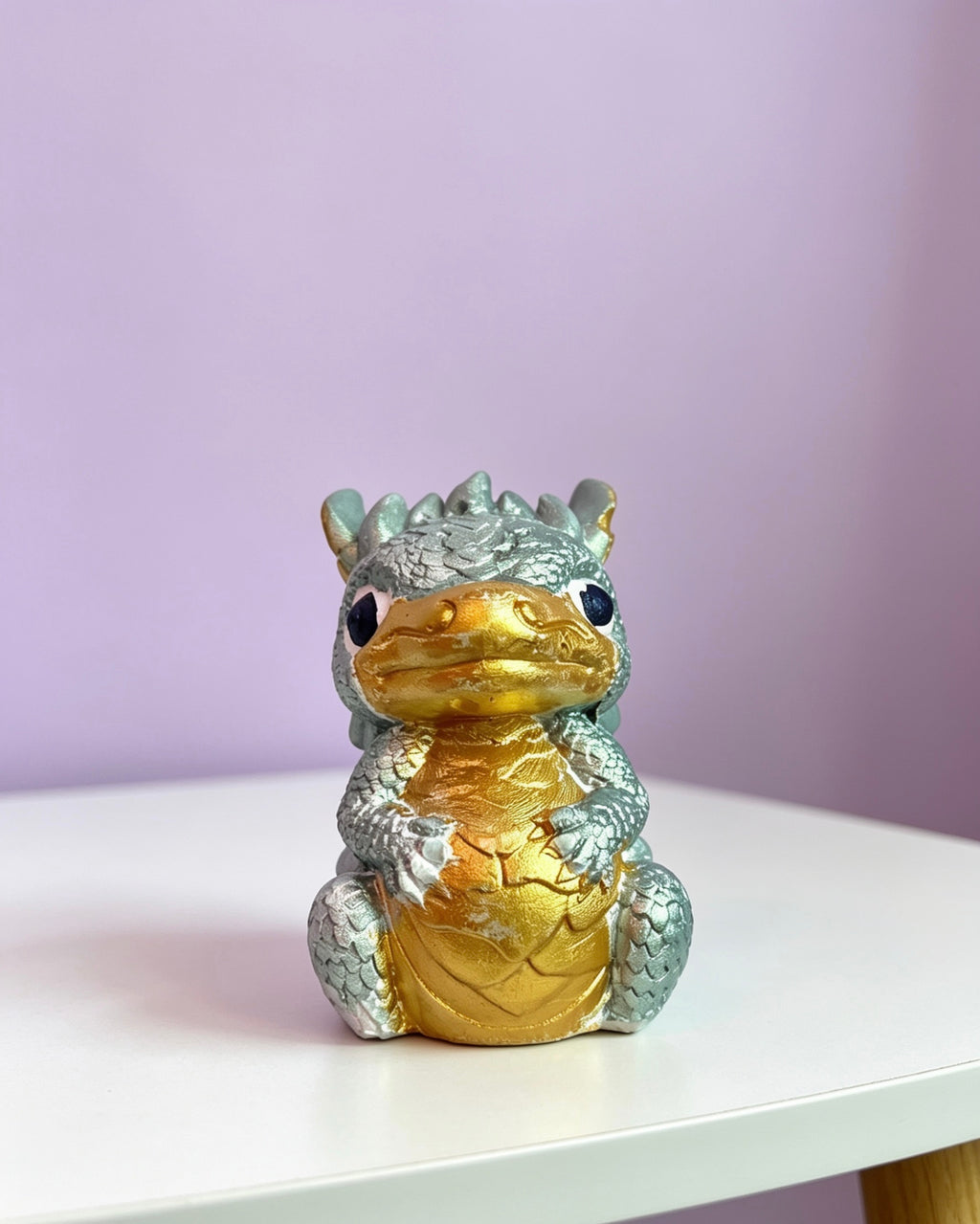 A small dragon figurine with a metallic gold belly and head, and silver-gray scaled body and limbs, sitting on a white tabletop against a soft lavender background.