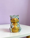 A small dragon figurine with a metallic gold belly and head, and silver-gray scaled body and limbs, sitting on a white tabletop against a soft lavender background.